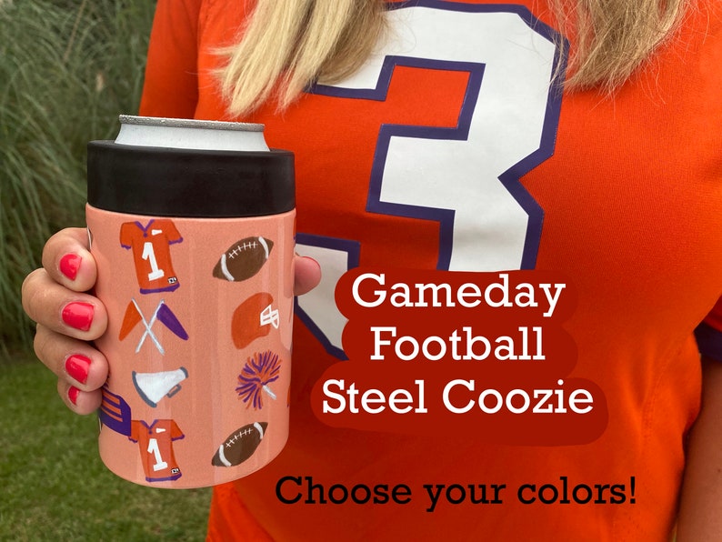 以下が含まれることがあります： サッカーをテーマにしたピンクとオレンジの断熱缶クーラー。クーラーには、サッカーボール、ジャージ、旗、メガホン、ポンポンが付いています。画像には、「Gameday Football Steel Coozie」と「Choose your colors!」のテキストが印刷されています。