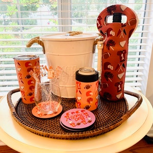 以下が含まれることがあります： ロープハンドルが付いた白いクーラー、ピンクとオレンジの断熱飲料ホルダー、ピンクとオレンジの断熱ワインボトルホルダー、透明なグラス、そして「ゲームデー」と書かれたピンクとオレンジのコースターが4つ。すべてのアイテムは茶色の籐製のトレイに乗っています。