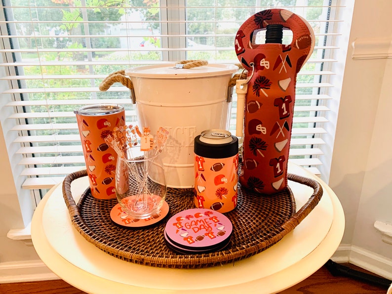 以下が含まれることがあります： ロープハンドルが付いた白いクーラー、ピンクとオレンジの断熱飲料ホルダー、ピンクとオレンジの断熱ワインボトルホルダー、透明なグラス、そして「ゲームデー」と書かれたピンクとオレンジのコースターが4つ。すべてのアイテムは茶色の籐製のトレイに乗っています。