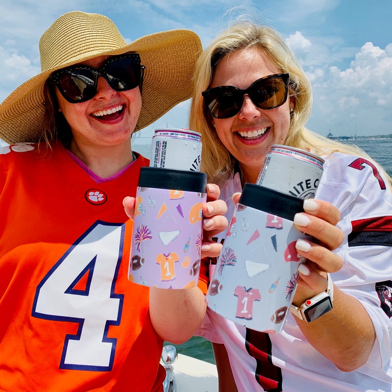 以下が含まれることがあります： 帽子をかぶった2人が、お祝いのデザインが施されたカラフルな缶クーラーを持っています。クーラーは、花火、紙吹雪、スポーツジャージなど、さまざまなパターンで飾られています。缶クーラーは、パーティーやイベントで飲み物を冷たく保つ、楽しくてお祝いの気分になれる方法です。