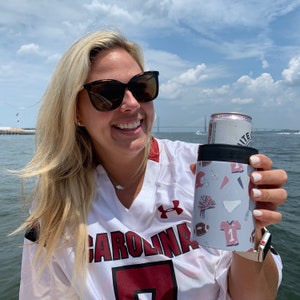 以下が含まれることがあります： 白いジャージを着て、「CAROLINA」と赤い数字の7が書かれた女性が、白い背景にカラフルなデザインが施された缶クーラーを持っています。缶クーラーの中には、側面に「White Claw」と書かれた白い缶が入っています。