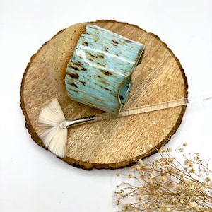 Puede incluir: Una taza de cerámica turquesa con motas marrones, colocada sobre una rodaja de madera. Un pincel con un cabezal blanco en forma de abanico y un mango transparente está al lado de la taza. Flores secas en la esquina inferior derecha.