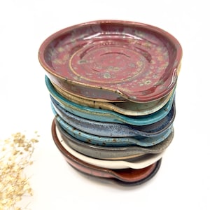 May include: A stack of eight handmade ceramic spoon rests in various colors, including red, brown, beige, blue, and gray. The spoon rests are all slightly different shapes and sizes, but they all have a similar curved design.