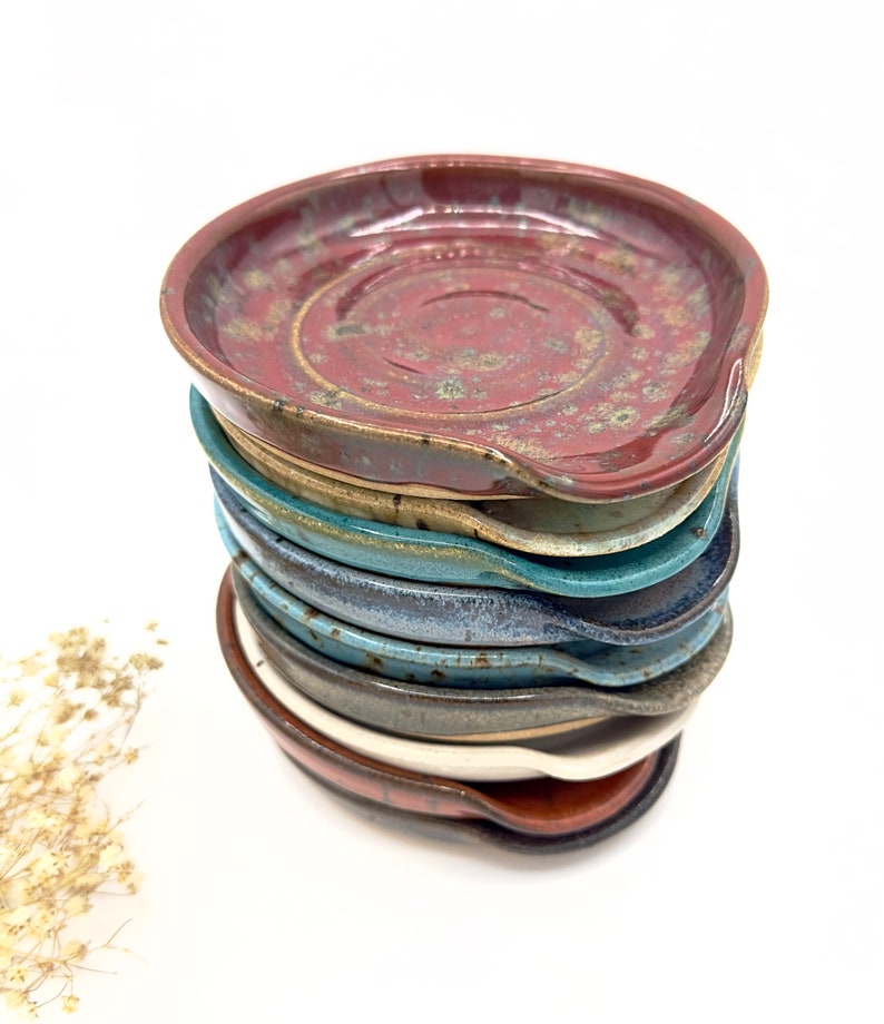 May include: A stack of eight handmade ceramic spoon rests in various colors, including red, brown, beige, blue, and gray. The spoon rests are all slightly different shapes and sizes, but they all have a similar curved design.