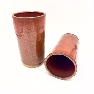 May include: Two cylindrical ceramic tumblers featuring a glossy, speckled reddish-brown glaze. One tumbler stands upright while the other lies on its side, revealing a smooth, dark interior. The base of each vessel is a contrasting light beige tone.