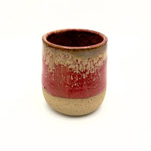 May include: A cylindrical ceramic cup featuring a speckled beige base and a vibrant red upper section. The rim displays a textured, dripped glaze effect where the red meets a brownish-tan tone, creating a distinct contrast against the raw clay bottom.