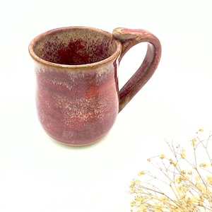May include: A handmade ceramic mug with a deep red glaze and a textured surface. The mug has a rounded body, a large handle, and a light brown rim. The mug is approximately 10 cm tall and is suitable for hot or cold beverages.
