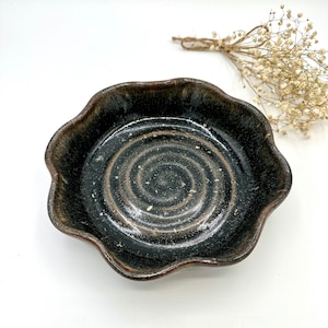 May include: A handmade ceramic bowl with a dark brown and black glaze. The bowl has a wavy, scalloped edge and a spiral design in the center. The bowl is approximately 6 inches in diameter.