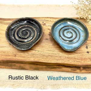 May include: Two handmade ceramic spoon rests, one in a rustic black glaze with a spiral design and the other in a weathered blue glaze with a spiral design. The spoon rests are displayed on a wooden surface.