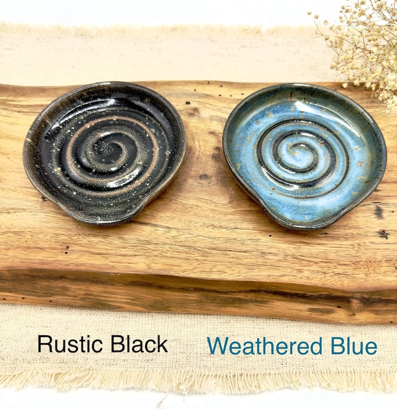 May include: Two handmade ceramic spoon rests, one in a rustic black glaze with a spiral design and the other in a weathered blue glaze with a spiral design. The spoon rests are displayed on a wooden surface.