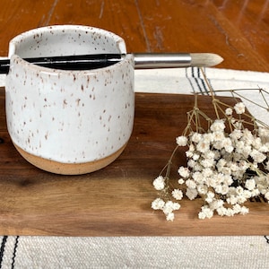 May include: White ceramic brush holder with brown speckles, a paintbrush resting on the rim, and a wooden cutting board with dried white flowers.
