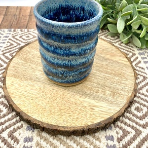 May include: A handmade ceramic tumbler with a textured, ribbed design. The cup features a gradient of blue hues, from a deep indigo to a lighter, almost white, shade. The cup is approximately 4 inches tall and sits on a wooden coaster.
