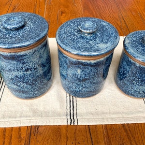 May include: Three blue ceramic storage jars with lids. The jars have a speckled blue glaze with a brown rim and lid. They are arranged on a white and black striped runner on a wooden surface. The jars are different sizes.