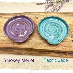 May include: Two ceramic spoon rests, one in a purple and brown swirl pattern labeled "Smokey Merlot" and the other in a blue and green swirl pattern labeled "Pacific Jade".