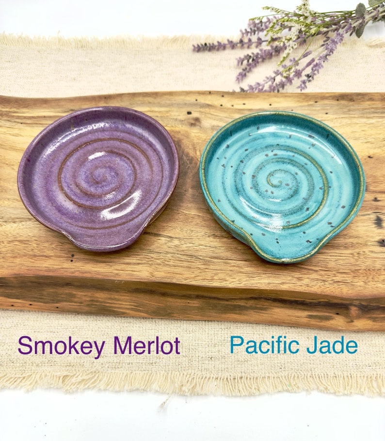 May include: Two ceramic spoon rests, one in a purple and brown swirl pattern labeled "Smokey Merlot" and the other in a blue and green swirl pattern labeled "Pacific Jade".