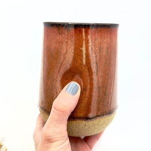 Könnte beinhalten: Eine handgefertigte Keramiktasse mit einer satten, rotbraunen Glasur. Die Tasse hat eine zylindrische Form mit einem leicht strukturierten, unglasierten Boden. Der Rand ist dunkelbraun, und die Tasse wird von einer Hand mit hellblauem Nagellack gehalten.