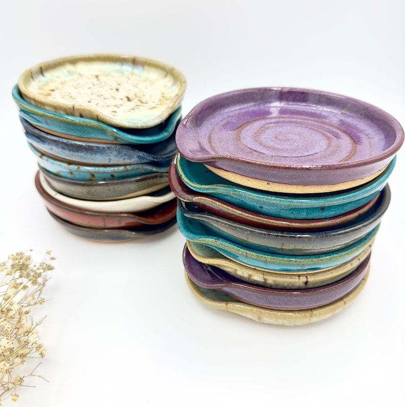 May include: A stack of handmade ceramic spoon rests in various shades of blue, purple, brown, and beige. The spoon rests are glazed and have a slightly curved shape.