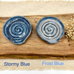 May include: Two ceramic spoon rests with a spiral design. The spoon rest on the left is a dark blue color with white speckles and is labeled "Stormy Blue". The spoon rest on the right is a light blue color with white speckles and is labeled "Frost Blue".