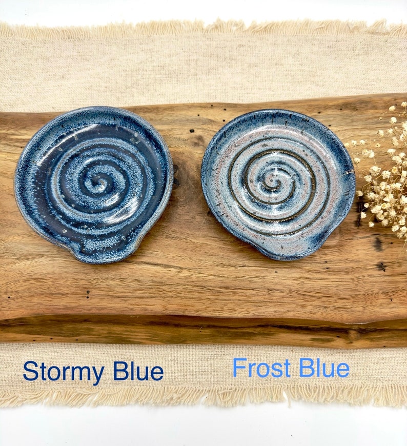 May include: Two ceramic spoon rests with a spiral design. The spoon rest on the left is a dark blue color with white speckles and is labeled "Stormy Blue". The spoon rest on the right is a light blue color with white speckles and is labeled "Frost Blue".