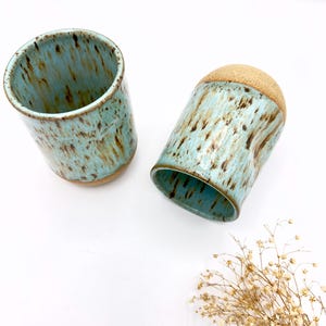 Könnte beinhalten: Zwei Keramikbecher mit hellblauer Glasur und braunen Flecken. Die Becher haben ein rustikales, handgemachtes Aussehen.