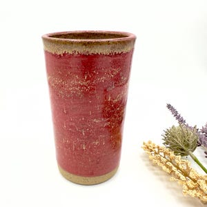 May include: A tall, cylindrical ceramic tumbler featuring a vibrant red glaze with a textured, speckled finish. The rim and the base are left in a natural, unglazed beige clay tone, creating a distinct contrast against the bright red body.