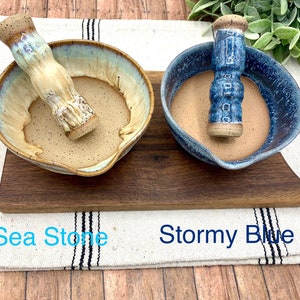 K&ouml;nnte beinhalten: Zwei M&ouml;rser- und St&ouml;&szlig;el-Sets aus Keramik. Das linke Set, beschriftet mit Sea Stone, zeigt eine cremefarbene und hellblaue glasierte Schale mit gesprenkeltem Inneren und passendem St&ouml;&szlig;el. Das rechte Set, Stormy Blue, hat eine dunkelblaue gesprenkelte Schale.