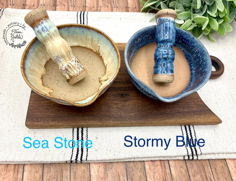 K&ouml;nnte beinhalten: Zwei M&ouml;rser- und St&ouml;&szlig;el-Sets aus Keramik. Das linke Set, beschriftet mit Sea Stone, zeigt eine cremefarbene und hellblaue glasierte Schale mit gesprenkeltem Inneren und passendem St&ouml;&szlig;el. Das rechte Set, Stormy Blue, hat eine dunkelblaue gesprenkelte Schale.
