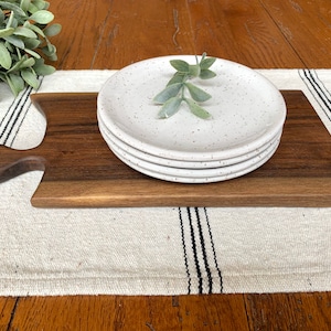 May include: A stack of four white speckled ceramic plates rests on a dark brown wooden serving board with a handle. The board is placed on a cream-colored striped table runner, with a sprig of greenery on top of the plates.