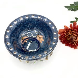 Puede incluir: Un cuenco de cerámica azul oscuro con un esmalte moteado, con una serie de agujeros espaciados uniformemente alrededor del borde. El cuenco contiene joyas, incluyendo pendientes y una pulsera azul. Una flor de color rojo-naranja está en el fondo.