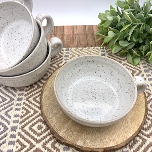 May include: Set of four white ceramic bowls with speckled brown glaze. The bowls have a rounded shape and a single handle. The bowls are arranged in a stack on a wooden surface.