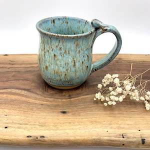 Può includere: Tazza in ceramica fatta a mano con un design unico del manico. La tazza presenta una smaltatura blu e marrone macchiettata, con una capacità di circa 350 ml. È esposta su una superficie di legno, mostrando il suo fascino rustico.