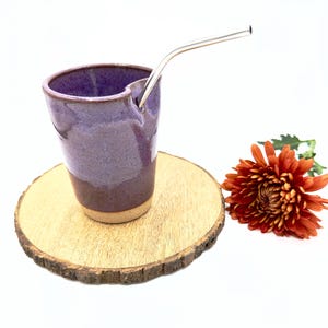 May include: A purple ceramic mug with a built-in straw holder and a stainless steel straw. The mug is sitting on a wooden coaster.