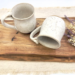 May include: Two white ceramic mugs with speckled brown glaze. The mugs have rounded bodies and a handle on one side. The mugs are sitting on a wooden cutting board.