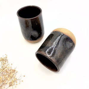 May include: Two ceramic mugs with a dark brown glaze and a light brown rim. The mugs are handmade and have a rustic look.