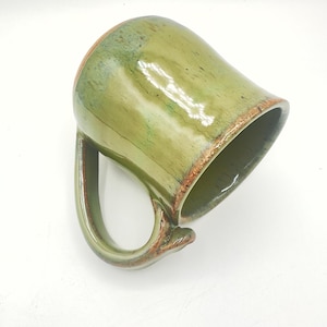 May include: A handmade ceramic mug with a unique, curved handle. The mug features a glossy green glaze with brown accents around the rim and handle base. The mug's interior is a darker shade of green.