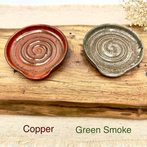 May include: Two ceramic spoon rests, one in a copper glaze and the other in a green smoke glaze. Both have a spiral design.