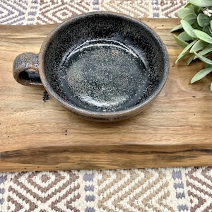 May include: A black ceramic bowl with a speckled glaze and a handle. The bowl is sitting on a wooden surface.