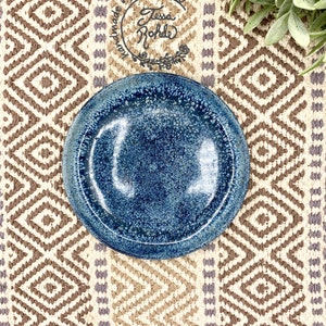 May include: A round ceramic plate with a deep blue speckled glaze and a smooth, glossy finish. The plate features a distinct rim and a dark, mottled texture throughout the surface. A circular stamp reads handmade pottery by Tessa Rohde.