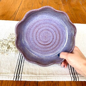 Puede incluir: Un cuenco de cerámica hecho a mano con un borde ondulado. El cuenco tiene una mezcla de tonos morados y lavanda, con un patrón en espiral en el centro. El cuenco está sostenido por una mano.