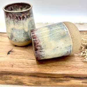 May include: Two ceramic mugs with a white glaze and brown and red accents. The mugs are sitting on a wooden surface.