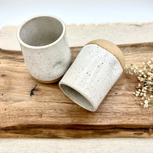 May include: Two white ceramic mugs with speckled brown glaze. The mugs are sitting on a wooden surface.