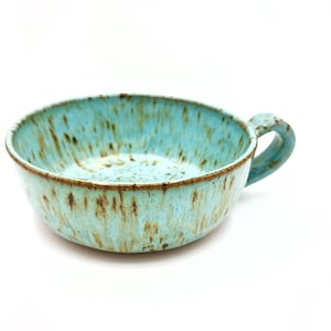 May include: A wide, shallow ceramic bowl with a single handle, featuring a speckled light blue glaze with brown vertical streaks and a dark brown rim. The interior and exterior surfaces display a mottled texture with visible color variations.