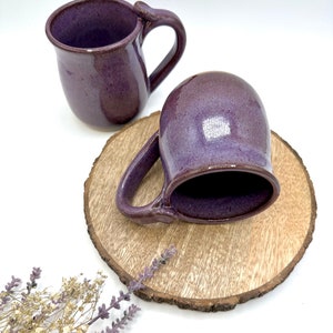 May include: Two handmade ceramic mugs in a deep purple glaze. The mugs have a rounded shape and a distinctive handle design. They are displayed on a wooden slice, with dried lavender sprigs in the foreground. The mugs are suitable for coffee or tea.