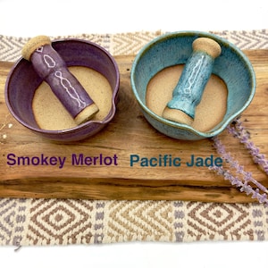 K&ouml;nnte beinhalten: Zwei M&ouml;rser- und St&ouml;&szlig;el-Sets aus Keramik. Ein Set ist in Smokey Merlot Violett glasiert, das andere in Pacific Jade Blau. Beide haben eine glatte, unglasierte beige Innenfl&auml;che und passende St&ouml;&szlig;el. Der Text Smokey Merlot und Pacific Jade ist sichtbar.