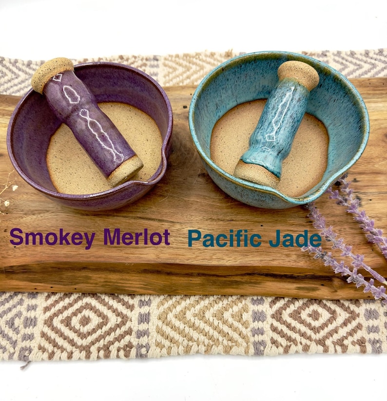 K&ouml;nnte beinhalten: Zwei M&ouml;rser- und St&ouml;&szlig;el-Sets aus Keramik. Ein Set ist in Smokey Merlot Violett glasiert, das andere in Pacific Jade Blau. Beide haben eine glatte, unglasierte beige Innenfl&auml;che und passende St&ouml;&szlig;el. Der Text Smokey Merlot und Pacific Jade ist sichtbar.