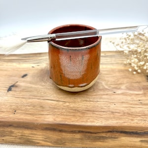 Puede incluir: Una taza de cerámica hecha a mano con un esmalte marrón moteado y una base color crema. Un pincel con mango transparente descansa dentro de la taza. La taza se exhibe sobre una superficie de madera, lo que sugiere un tema de cerámica o arte.