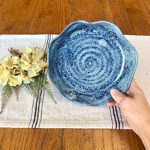Puede incluir: Un plato de cerámica azul hecho a mano con un borde ondulado único. El plato presenta un diseño en espiral moteado en tonos de azul y blanco. El plato se sostiene sobre un camino de mesa de lino a rayas sobre una mesa de madera.