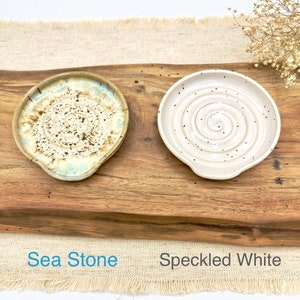 May include: Two ceramic spoon rests, one with a speckled white glaze and the other with a sea-stone glaze. The spoon rests are on a wooden surface with the words "Sea Stone" and "Speckled White" written below them.