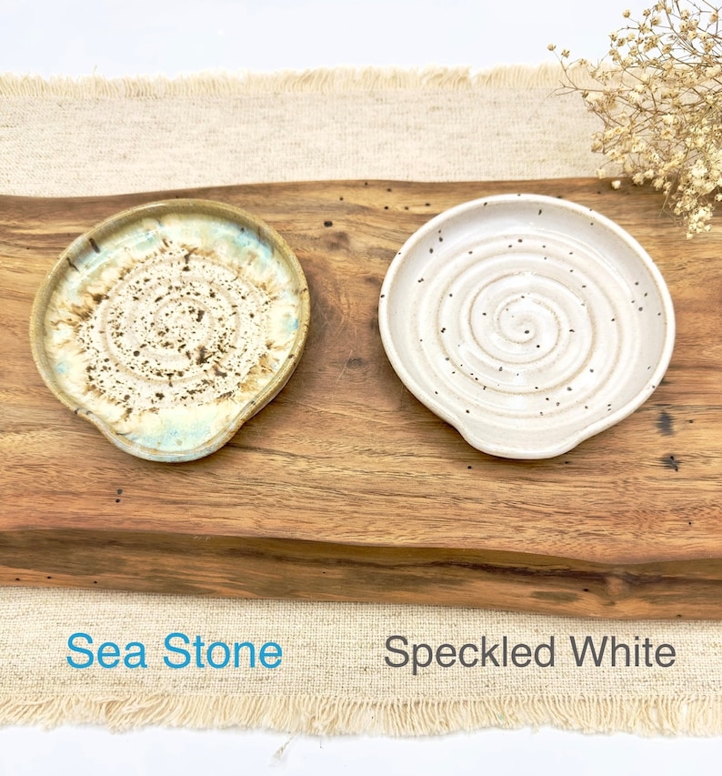 May include: Two ceramic spoon rests, one with a speckled white glaze and the other with a sea-stone glaze. The spoon rests are on a wooden surface with the words "Sea Stone" and "Speckled White" written below them.