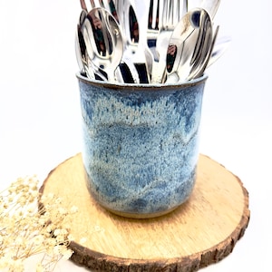 May include: A blue and white ceramic utensil holder filled with silverware, including forks, spoons, and knives. The holder sits on a round wooden slice with dried flowers on the side. The holder has a speckled glaze.
